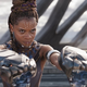 Letitia Wright in action as Shuri in Black Panther, raising two panther-shaped gauntlets up to fire.